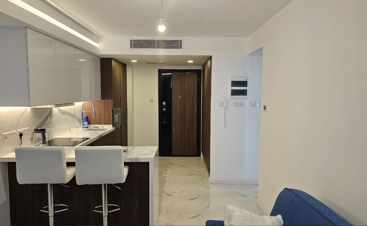 kıbrıs Küçük bir mutfak, mermer tezgahta iki beyaz bar taburesi, ahşap dolaplar, beyaz karo zemin ve sağda kısmen görünen mavi bir kanepe ile modern daire içi. Arka planda bir koridor ve giriş kapısı var. aysan_giti