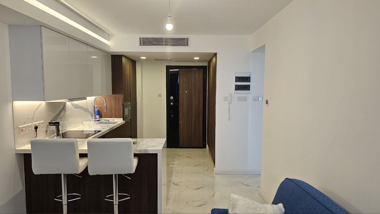 kıbrıs Küçük bir mutfak, mermer tezgahta iki beyaz bar taburesi, ahşap dolaplar, beyaz karo zemin ve sağda kısmen görünen mavi bir kanepe ile modern daire içi. Arka planda bir koridor ve giriş kapısı var. aysan_giti