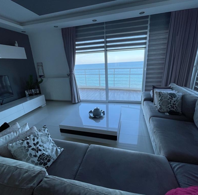 kıbrıs Gri kanepeler, beyaz bir sehpa, düz ekran TV ve okyanusa bakan geniş pencerelere sahip modern oturma odası, muhteşem manzaraya ve doğal ışığa sahip bir Kuzey Kıbrıs satılık daire arayanlar için idealdir. aysan_giti