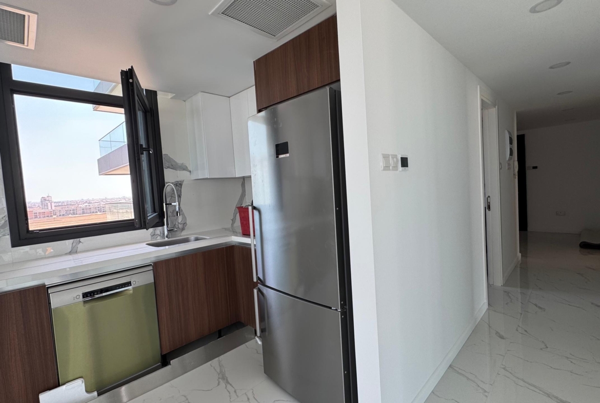 kıbrıs Grand Sapphire Resort & Residence'ın 7 yıldızlı otel konseptinde yaşam Kıbrıs sunan ünitesinde kahverengi dolaplar, paslanmaz çelik buzdolabı ve mermer tezgahlı modern mutfak. Lavabonun üzerindeki pencereden güneş ışığı süzülüyor. aysan_giti