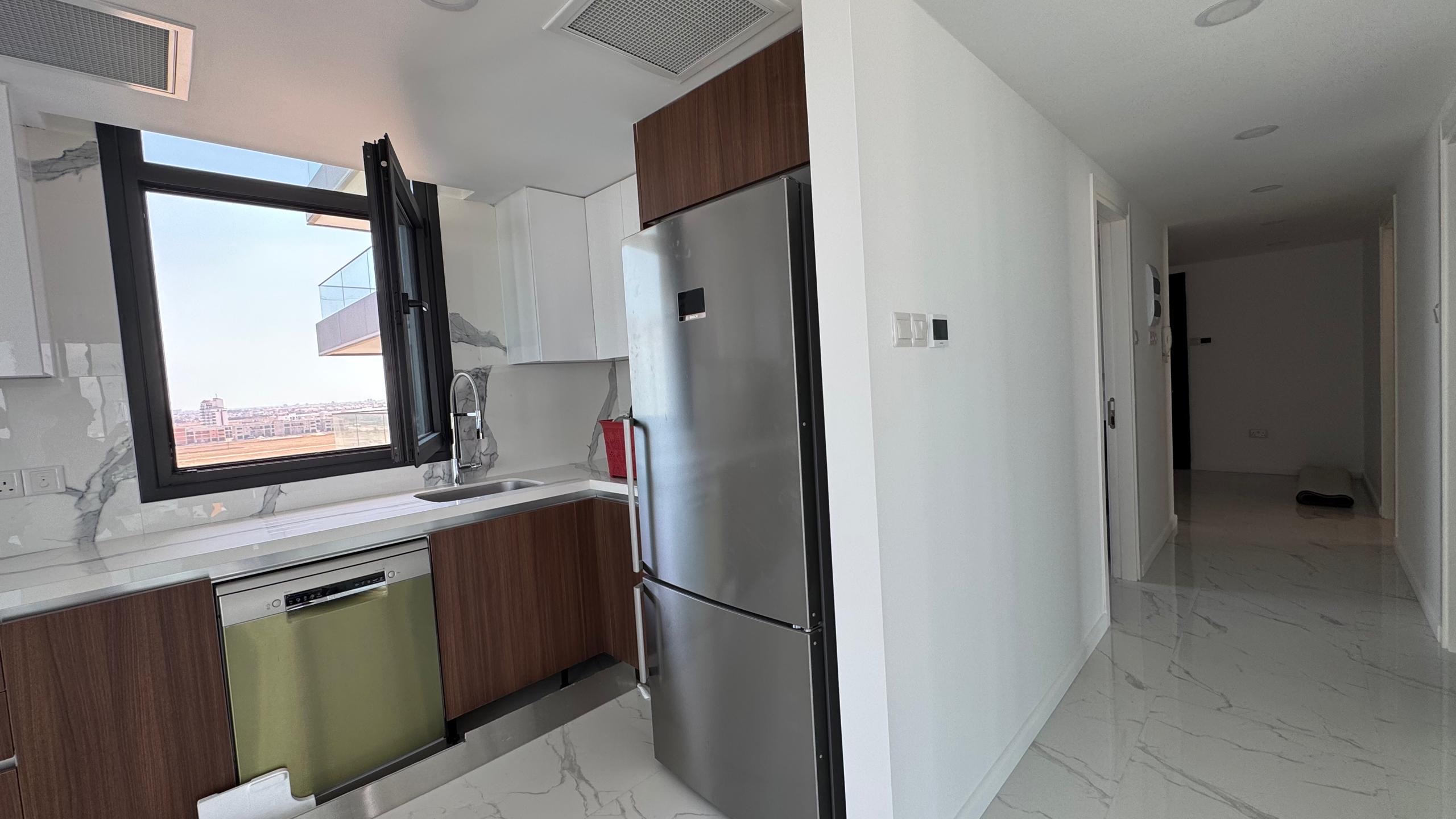 kıbrıs Grand Sapphire Resort & Residence'ın 7 yıldızlı otel konseptinde yaşam Kıbrıs sunan ünitesinde kahverengi dolaplar, paslanmaz çelik buzdolabı ve mermer tezgahlı modern mutfak. Lavabonun üzerindeki pencereden güneş ışığı süzülüyor. aysan_giti