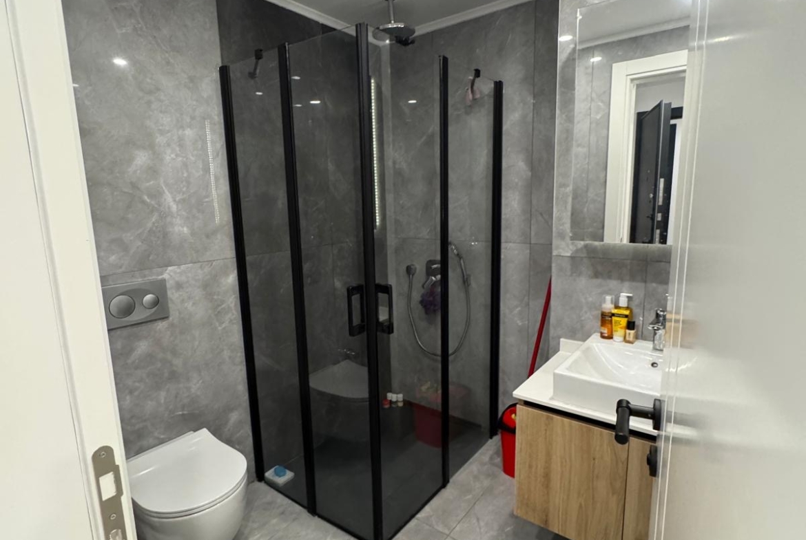 kıbrıs Gri mermer duvarlar ve zemin, siyah çerçeveli cam duş kabini, duvara monte tuvalet, beyaz lavabolu ahşap makyaj masası, ayna, banyo malzemeleri ve yerde yuvarlak bir halı bulunan modern banyo. aysan_giti