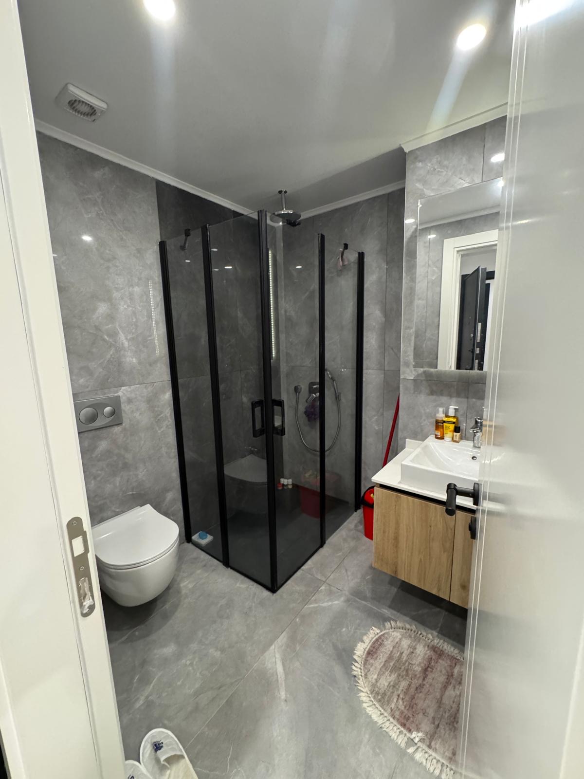 kıbrıs Gri mermer duvarlar ve zemin, siyah çerçeveli cam duş kabini, duvara monte tuvalet, beyaz lavabolu ahşap makyaj masası, ayna, banyo malzemeleri ve yerde yuvarlak bir halı bulunan modern banyo. aysan_giti kıbrıs Gri mermer duvarlar ve zemin, siyah çerçeveli cam duş kabini, duvara monte tuvalet, beyaz lavabolu ahşap makyaj masası, ayna, banyo malzemeleri ve yerde yuvarlak bir halı bulunan modern banyo. aysan_giti