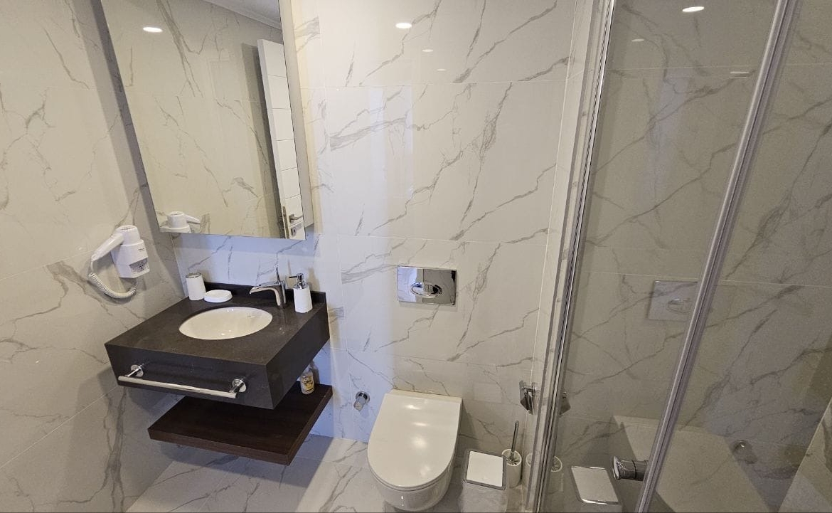 kıbrıs Beyaz mermer duvarlar, banyo malzemeleri içeren yüzer lavabo, duvara monte saç kurutma makinesi, ayna, tuvalet ve sağda cam duş kabini bulunan modern banyo. aysan_giti