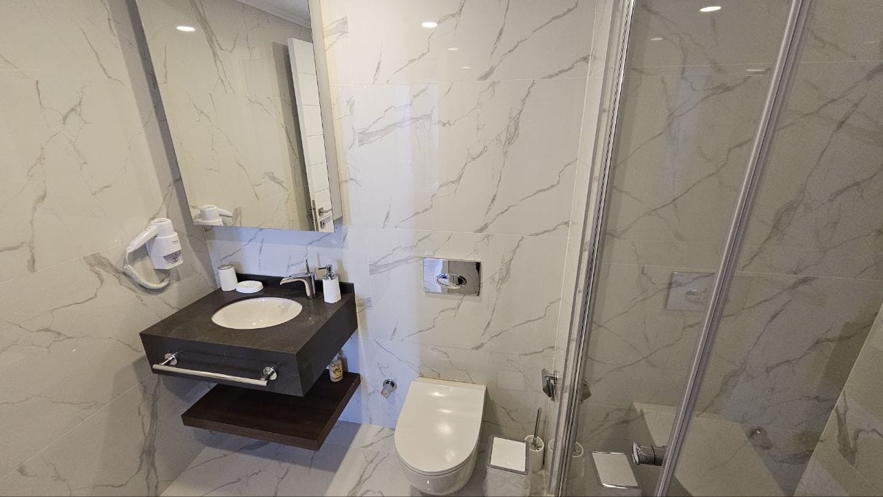 kıbrıs Beyaz mermer duvarlar, banyo malzemeleri içeren yüzer lavabo, duvara monte saç kurutma makinesi, ayna, tuvalet ve sağda cam duş kabini bulunan modern banyo. aysan_giti