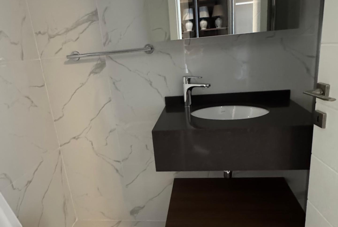 kıbrıs Grand Sapphire Resort & Residence Kıbrıs tarafından sunulan 7 yıldızlı otel konseptinde yaşam Kıbrıs'ı yansıtan beyaz mermer duvarlar ve zemin, duvara monte siyah lavabo, üstte ayna, solda tuvalet ve açık ahşap raflı modern banyo. aysan_giti