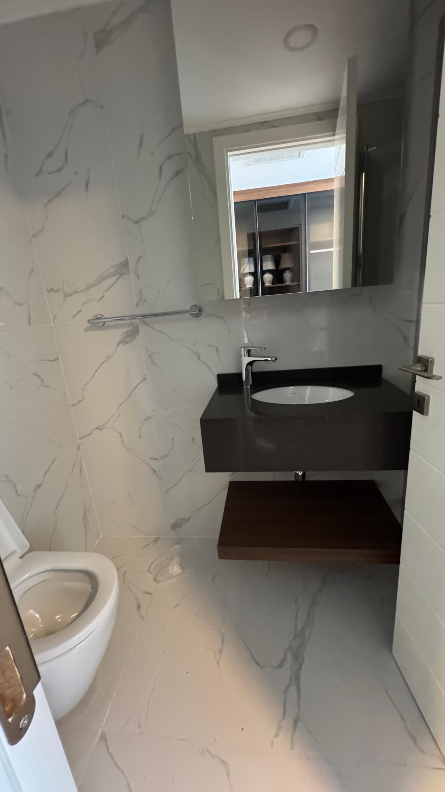 kıbrıs Grand Sapphire Resort & Residence Kıbrıs tarafından sunulan 7 yıldızlı otel konseptinde yaşam Kıbrıs'ı yansıtan beyaz mermer duvarlar ve zemin, duvara monte siyah lavabo, üstte ayna, solda tuvalet ve açık ahşap raflı modern banyo. aysan_giti