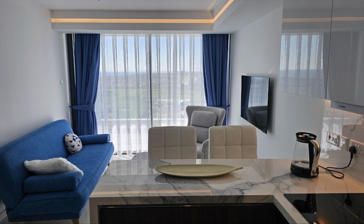 kıbrıs Mavi bir kanepe, koltuk ve mermer bir tezgahın yanında beyaz yemek sandalyeleri bulunan modern bir yaşam alanı. Mavi perdeli büyük pencereler dışarıdaki yeşil manzarayı gözler önüne seriyor. Tezgahın üzerinde bir cam su ısıtıcısı duruyor. aysan_giti