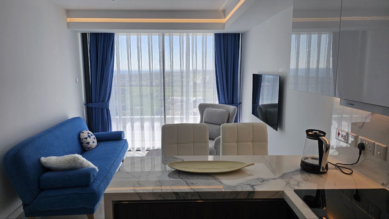 kıbrıs Mavi bir kanepe, koltuk ve mermer bir tezgahın yanında beyaz yemek sandalyeleri bulunan modern bir yaşam alanı. Mavi perdeli büyük pencereler dışarıdaki yeşil manzarayı gözler önüne seriyor. Tezgahın üzerinde bir cam su ısıtıcısı duruyor. aysan_giti