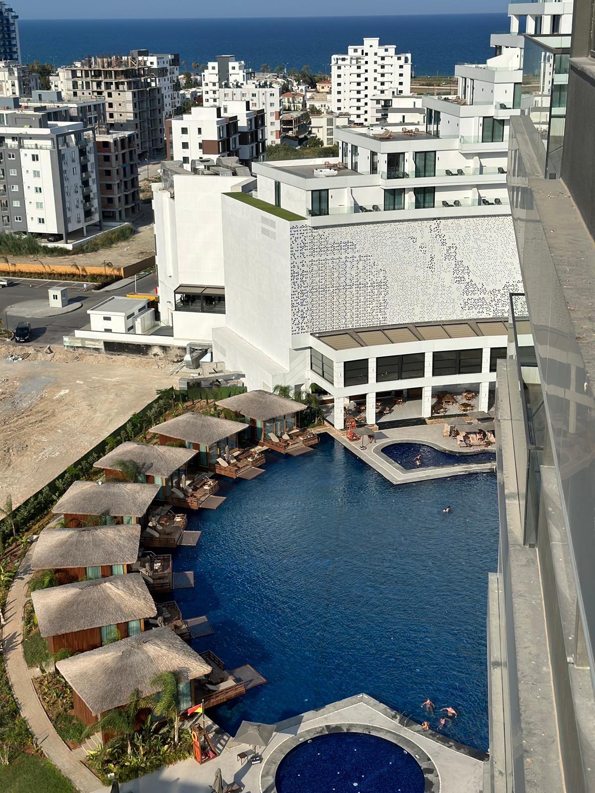 kıbrıs Yüksek beyaz binaların yanında sazdan kabanalar ve şezlonglar bulunan modern bir otelin havuz alanının havadan görünümü. Ötesinde okyanus görülüyor. İskele Long Beach satılık daire arayanlar veya Kuzey Kıbrıs gayrimenkulde Aysan Giti emlak'ı keşfetmek için mükemmeldir. aysan_giti