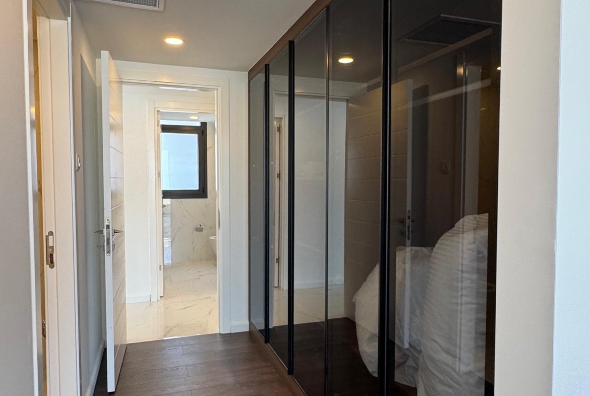 kıbrıs Koyu ahşap zeminli modern bir koridor, sağda siyah cam cepheli dolaplar, beyaz duvarlar, gömme aydınlatma ve aydınlık bir banyo manzarası - Aysan Giti emlak ile Kuzey Kıbrıs satılık daire seçenekleri arayanlar için ideal. aysan_giti