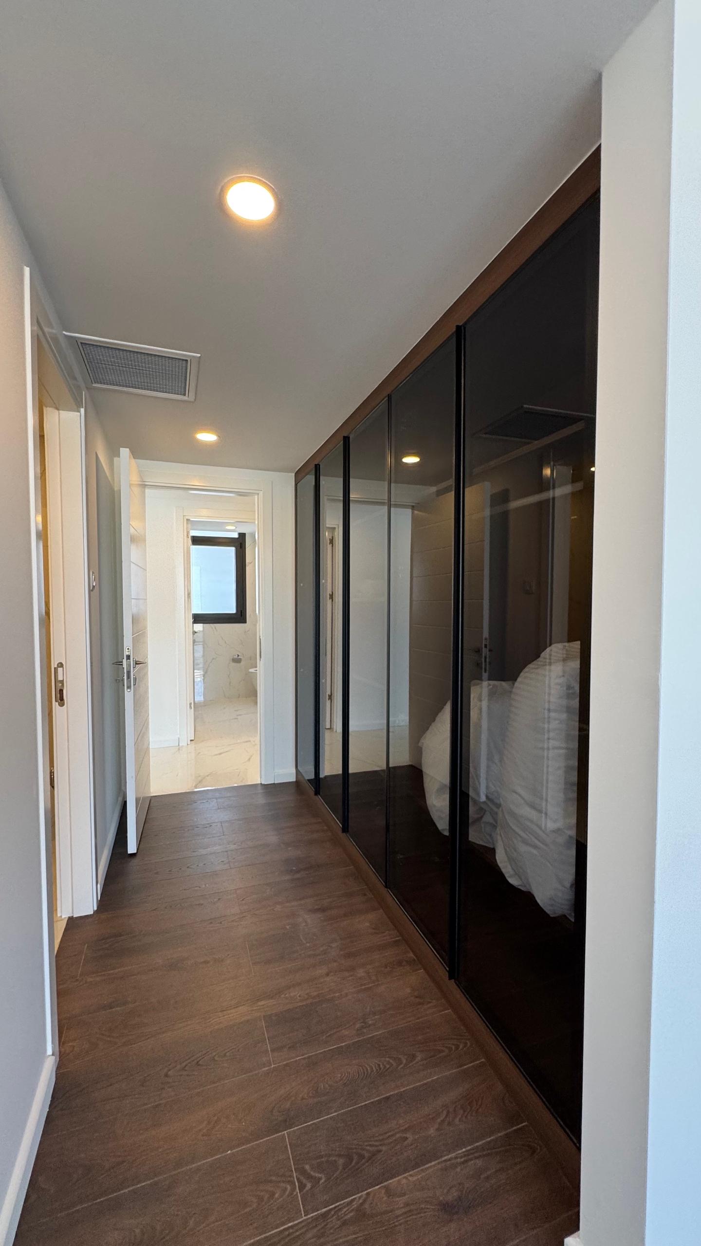 kıbrıs Koyu ahşap zeminli modern bir koridor, sağda siyah cam cepheli dolaplar, beyaz duvarlar, gömme aydınlatma ve aydınlık bir banyo manzarası - Aysan Giti emlak ile Kuzey Kıbrıs satılık daire seçenekleri arayanlar için ideal. aysan_giti
