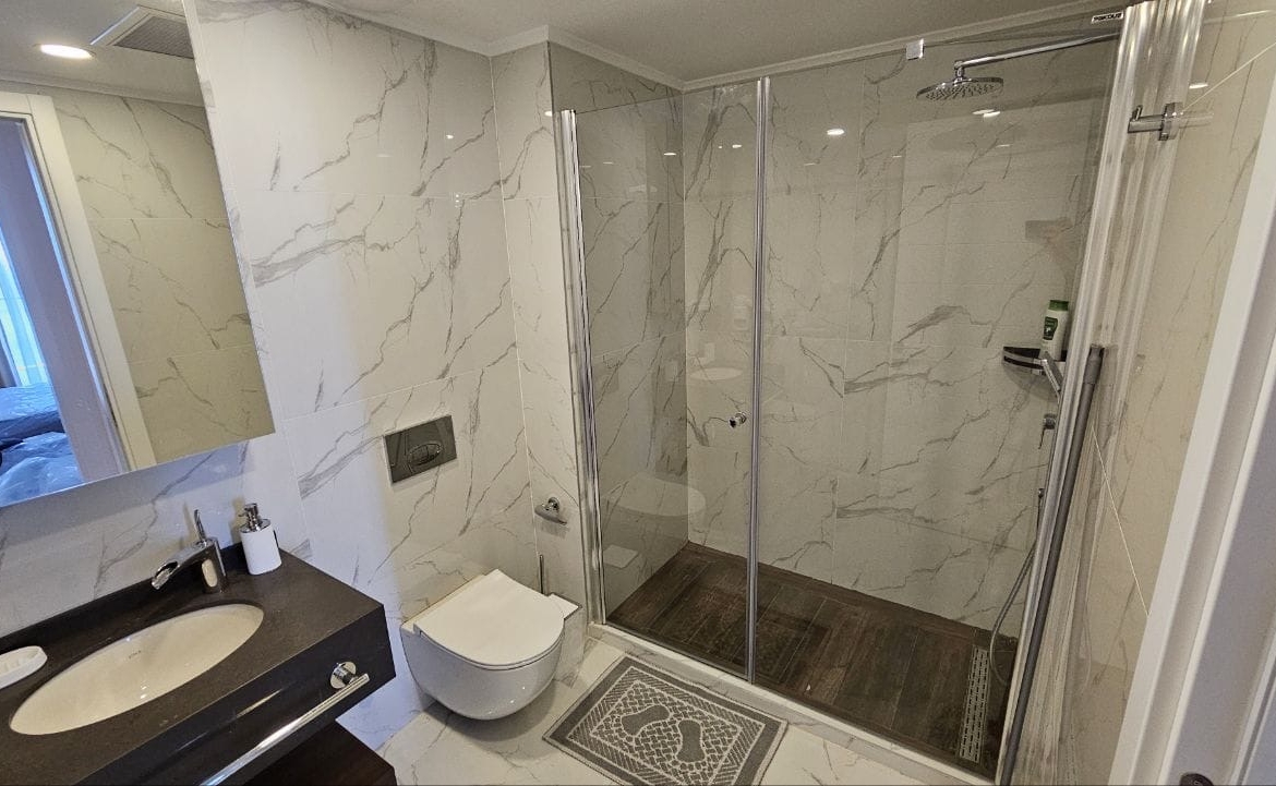 kıbrıs Beyaz mermer duvarlar, camla çevrili duş, duvara monte tuvalet, lavabo, ayna ve banyo malzemeleri içeren siyah bir tezgahtan oluşan modern bir banyo. Duşun içindeki zemin ahşap bir dokuya ve dışında küçük bir halıya sahiptir. aysan_giti