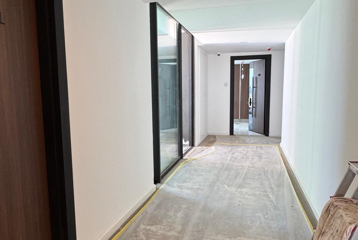 kıbrıs Beyaz duvarları, çıplak zemini ve büyük cam pencereleriyle inşaat halindeki bir koridor, modern Kuzey Kıbrıs gayrimenkulünün ipuçlarını veriyor. Kapılar solda sıralanıyor; bir merdiven ve kumaş sağda duruyor, güneş ışığı cam panellerden ve uzaktaki bir kapıdan akıyor. aysan_giti