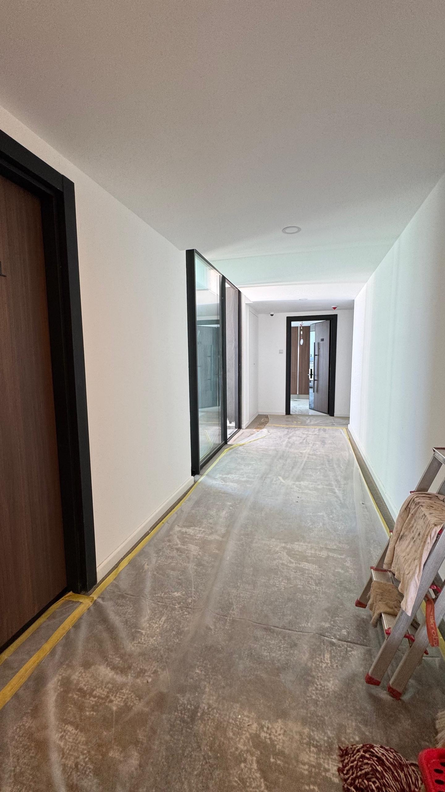 kıbrıs Beyaz duvarları, çıplak zemini ve büyük cam pencereleriyle inşaat halindeki bir koridor, modern Kuzey Kıbrıs gayrimenkulünün ipuçlarını veriyor. Kapılar solda sıralanıyor; bir merdiven ve kumaş sağda duruyor, güneş ışığı cam panellerden ve uzaktaki bir kapıdan akıyor. aysan_giti