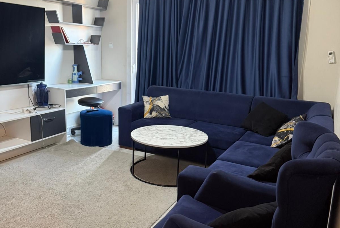 kıbrıs Büyük mavi kesitli kanepe, siyah ve sarı yastıklar, yuvarlak mermer sehpa, mavi perdeler, bej halı, duvara monte TV ve dekoratif eşyalarla dolu beyaz raflardan oluşan modern bir oturma odası. aysan_giti