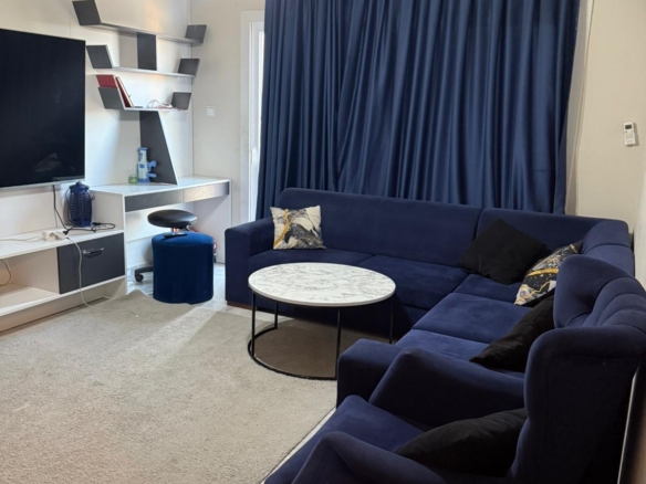 kıbrıs Büyük mavi kesitli kanepe, siyah ve sarı yastıklar, yuvarlak mermer sehpa, mavi perdeler, bej halı, duvara monte TV ve dekoratif eşyalarla dolu beyaz raflardan oluşan modern bir oturma odası. aysan_giti