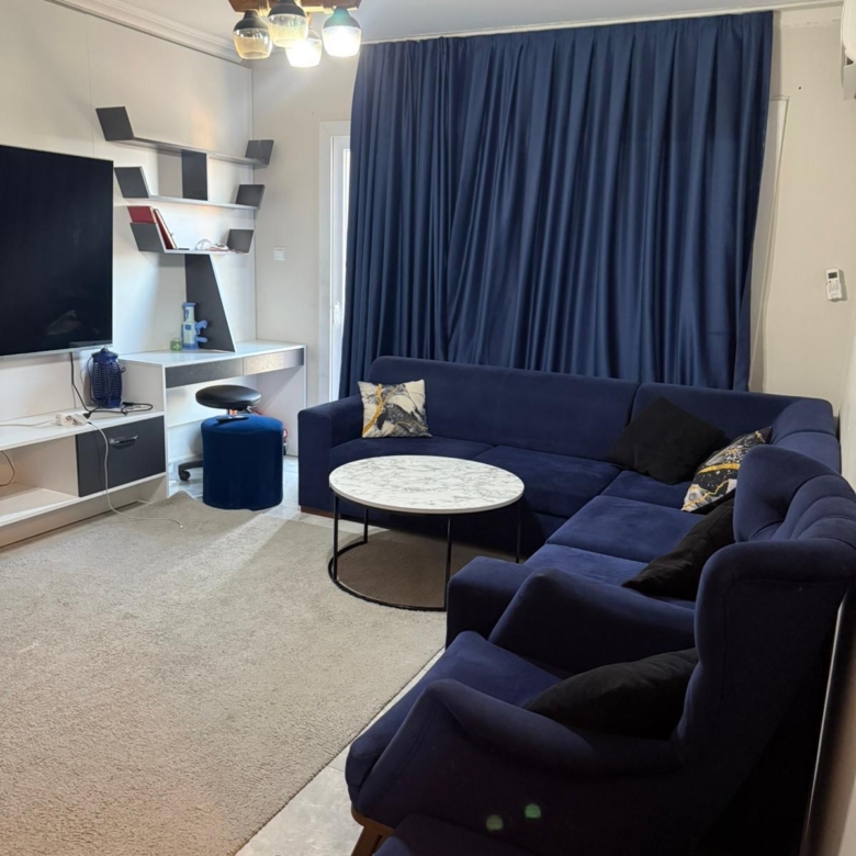 kıbrıs Büyük mavi kesitli kanepe, siyah ve sarı yastıklar, yuvarlak mermer sehpa, mavi perdeler, bej halı, duvara monte TV ve dekoratif eşyalarla dolu beyaz raflardan oluşan modern bir oturma odası. aysan_giti