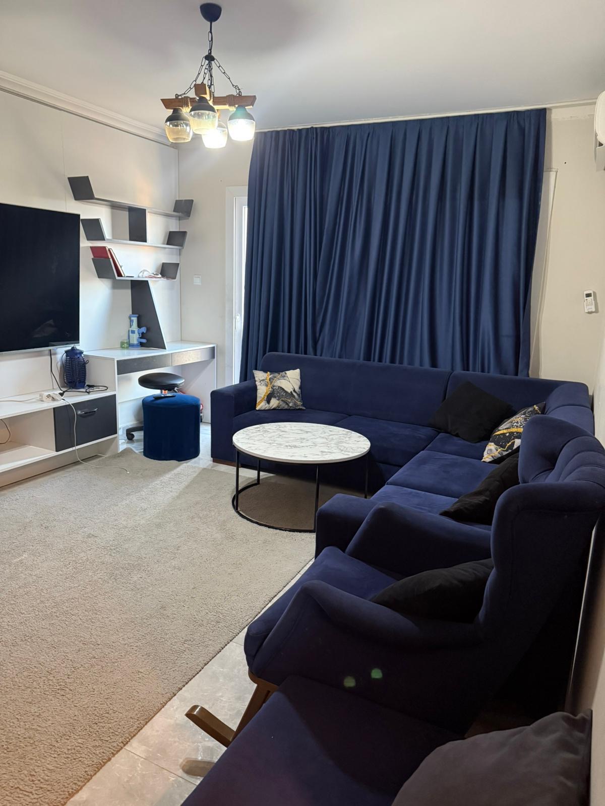 kıbrıs Büyük mavi kesitli kanepe, siyah ve sarı yastıklar, yuvarlak mermer sehpa, mavi perdeler, bej halı, duvara monte TV ve dekoratif eşyalarla dolu beyaz raflardan oluşan modern bir oturma odası. aysan_giti