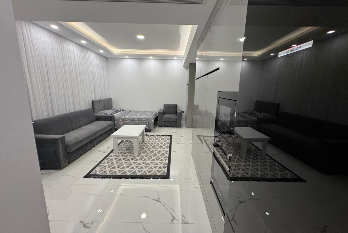 kıbrıs Gri kanepeler, beyaz bir sehpa, desenli halılar ve beyaz fayans döşemeli modern bir oturma odası. Odanın tavan ışıkları ve duvarları gömme olup bir tarafında mobilyaları yansıtan cam bir bölme bulunmaktadır. aysan_giti