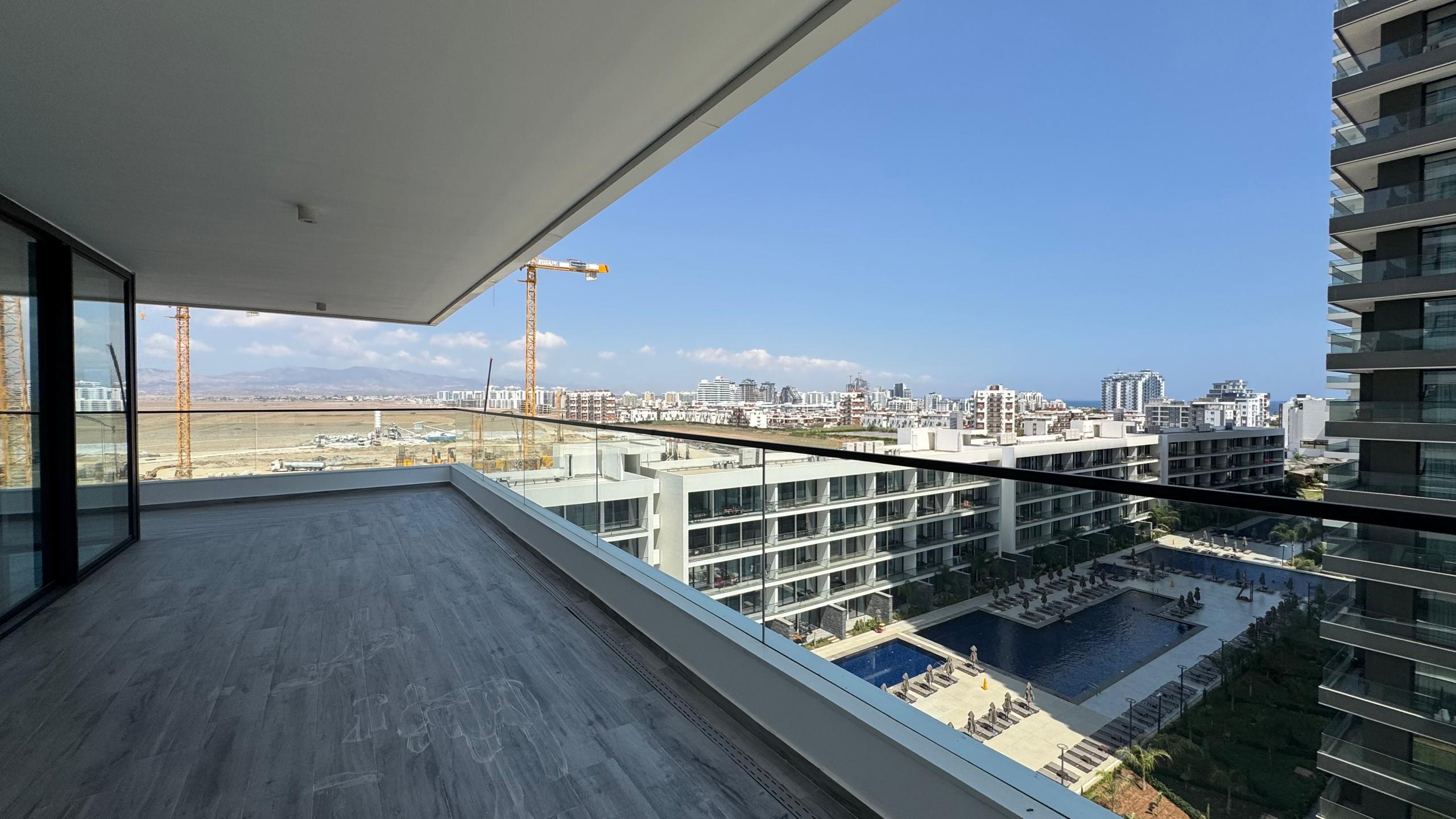 kıbrıs Modern apartmanlara, sonsuzluk havuzuna ve Grand Sapphire Resort & Residence kumarhanesine, inşaat vinçlerine ve masmavi bir gökyüzü altında uzaktaki şehir manzarasına bakan cam korkuluklu geniş bir balkondan görünüm. aysan_giti