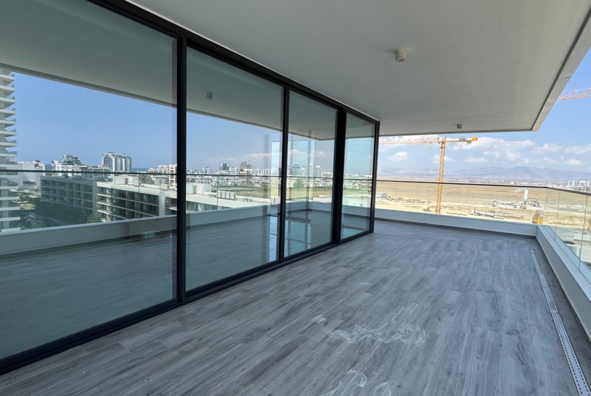 kıbrıs Grand Sapphire Resort & Residence Kıbrıs'ın cam korkuluklu ve geniş sürgülü kapılı ferah modern balkonu, masmavi gökyüzü altında yakındaki binaların, bir inşaat vincinin ve uzaktaki açık alanın manzarasını sunuyor. aysan_giti