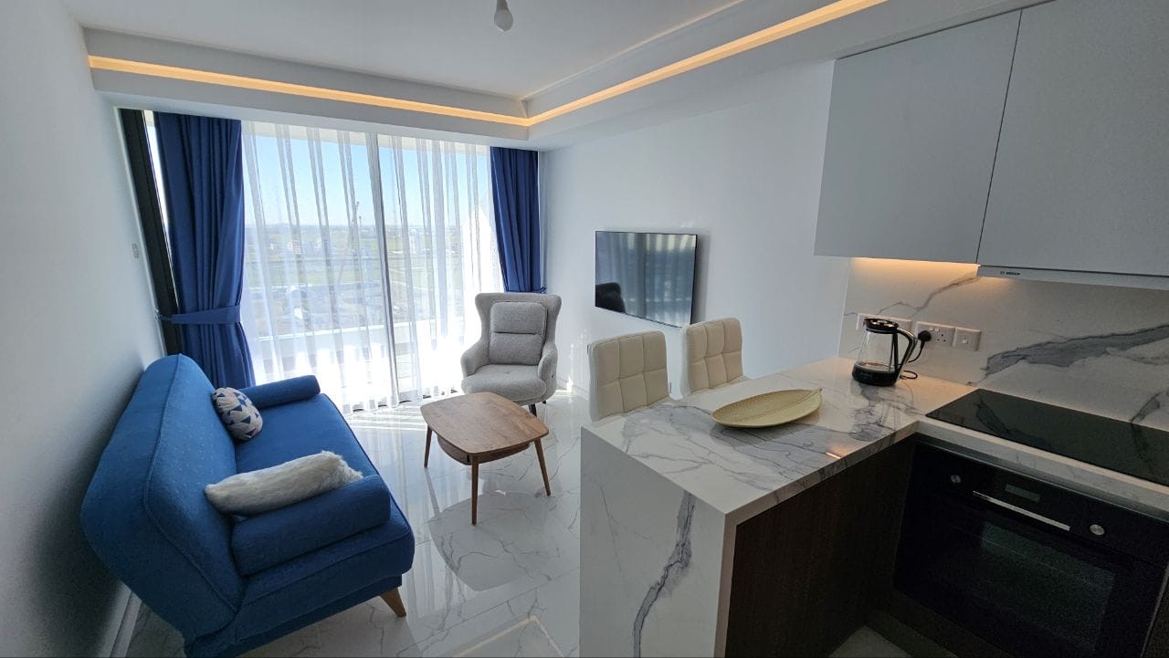 kıbrıs Mavi bir kanepe, koltuk, küçük ahşap masa, mermer tarzı tezgahlar, beyaz bar tabureleri, duvara monte TV ve doğal ışık alan mavi perdeli büyük pencereler içeren modern oturma odası ve mutfak. aysan_giti