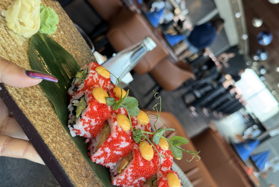 kıbrıs Bir el, yeşilliklerle süslenmiş, kırmızı karaca ve sarı sosla kaplı suşi rulolarının bulunduğu dikdörtgen bir taş tabağı tutuyor. Yanında zencefil ve wasabi var. Arka planda şık bir restoran bekliyor - Kuzey Kıbrıs satılık daire'yi gezdikten sonra mükemmel. aysan_giti