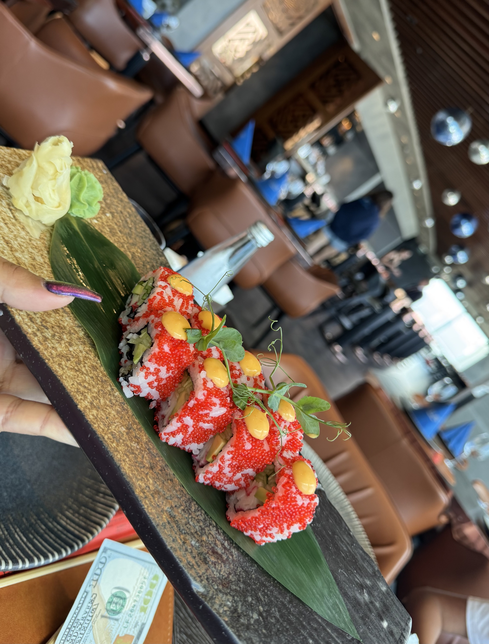 kıbrıs Bir el, yeşilliklerle süslenmiş, kırmızı karaca ve sarı sosla kaplı suşi rulolarının bulunduğu dikdörtgen bir taş tabağı tutuyor. Yanında zencefil ve wasabi var. Arka planda şık bir restoran bekliyor - Kuzey Kıbrıs satılık daire'yi gezdikten sonra mükemmel. aysan_giti