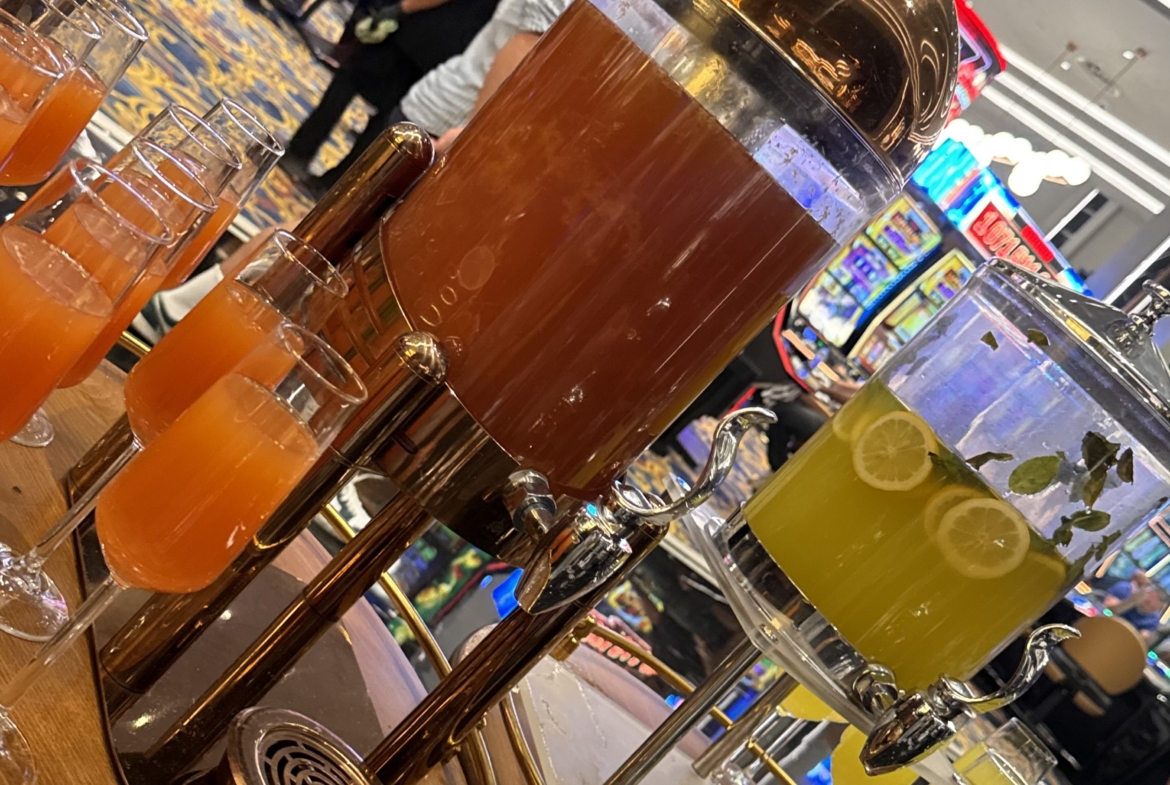 kıbrıs Kumar makinelerinin ve parlak ışıkların yakınındaki bir kumarhanede bulunan içecek istasyonunda turuncu ve sarı renkli iki büyük içecek dağıtıcısı ve bunların etrafına dizilmiş bardaklar bulunuyor. Nike tişörtlü bir adam yakınlarda duruyor - Kuzey Kıbrıs gayrimenkul seçeneklerini keşfeden konuklar için mükemmel. aysan_giti