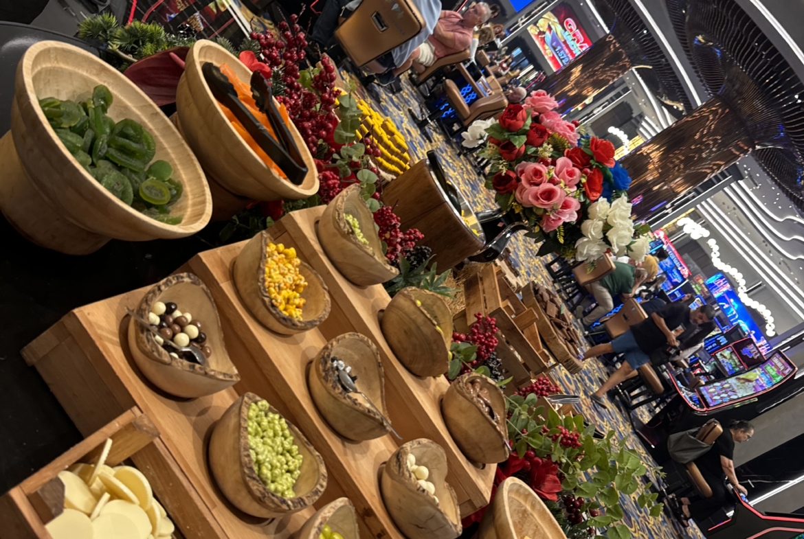 kıbrıs Ahşap kaselerde çeşitli atıştırmalıklar bulunan bir büfe masası, kalabalık bir kumarhane alanının önünde duruyor; çiçekler, insanlar ve kumar makineleri arka planı dolduruyor ve Kuzey Kıbrıs satılık daire ve canlı Kuzey Kıbrıs gayrimenkul sahnelerinin canlı atmosferini çağrıştırıyor. aysan_giti