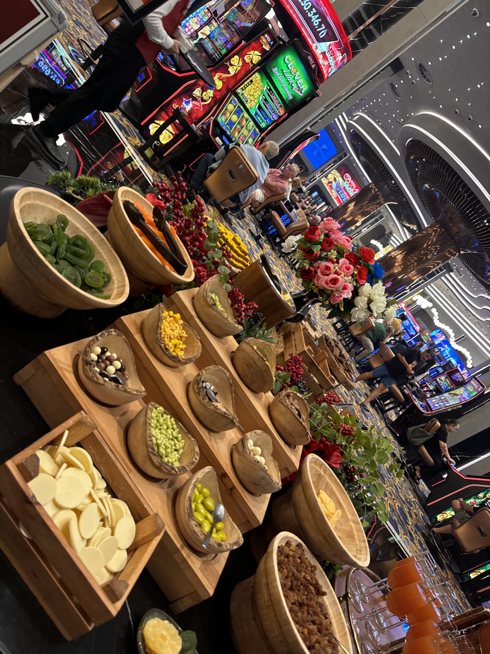 kıbrıs Ahşap kaselerde çeşitli atıştırmalıklar bulunan bir büfe masası, kalabalık bir kumarhane alanının önünde duruyor; çiçekler, insanlar ve kumar makineleri arka planı dolduruyor ve Kuzey Kıbrıs satılık daire ve canlı Kuzey Kıbrıs gayrimenkul sahnelerinin canlı atmosferini çağrıştırıyor. aysan_giti