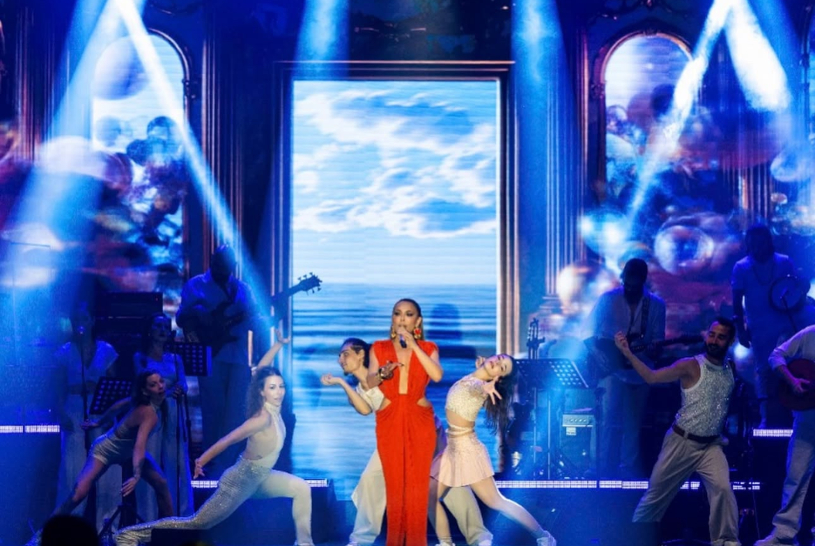 kıbrıs Kırmızı elbiseli bir kadın şarkıcı, dansçılar ve müzisyenler, dramatik ışıklar ve gökyüzünü gösteren video ekranları eşliğinde sahnede performans sergiliyor. Masalardaki izleyiciler İskele Long Beach satılık daire fırsatlarından sadece birkaç adım ötede gösterinin tadını çıkarıyor. aysan_giti