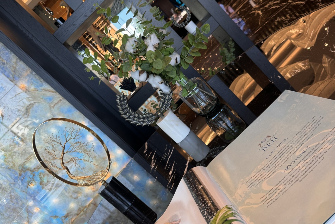 kıbrıs Parlak siyah zemini, zarif dekoru ve çiçekli vazosu olan kitaplığıyla şık bir salon; Kuzey Kıbrıs kiralık daire arayanlar için ideal. Masanın üzerinde açık bir sanat kitabı duruyor ve arka planda insanlar görünüyor. aysan_giti