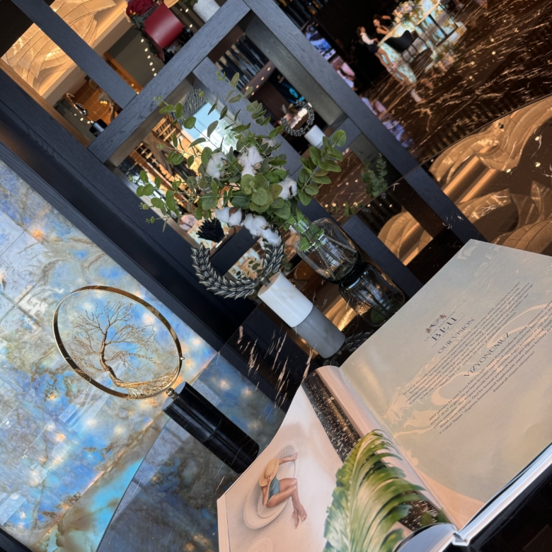 kıbrıs Parlak siyah zemini, zarif dekoru ve çiçekli vazosu olan kitaplığıyla şık bir salon; Kuzey Kıbrıs kiralık daire arayanlar için ideal. Masanın üzerinde açık bir sanat kitabı duruyor ve arka planda insanlar görünüyor. aysan_giti