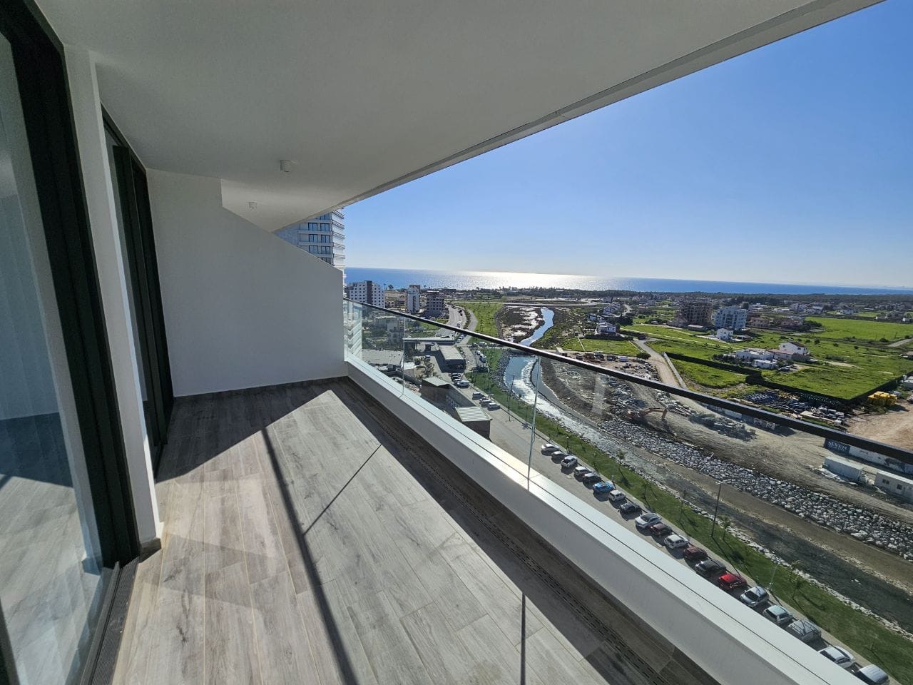kıbrıs Cam korkuluklu modern apartman balkonu, masmavi bir gökyüzünün altında kıyı şehir manzarasına, yeşil alanlara, binalara ve denize bakıyor. Güneş ışığı karo zemin üzerinde gölgeler oluşturuyor. aysan_giti