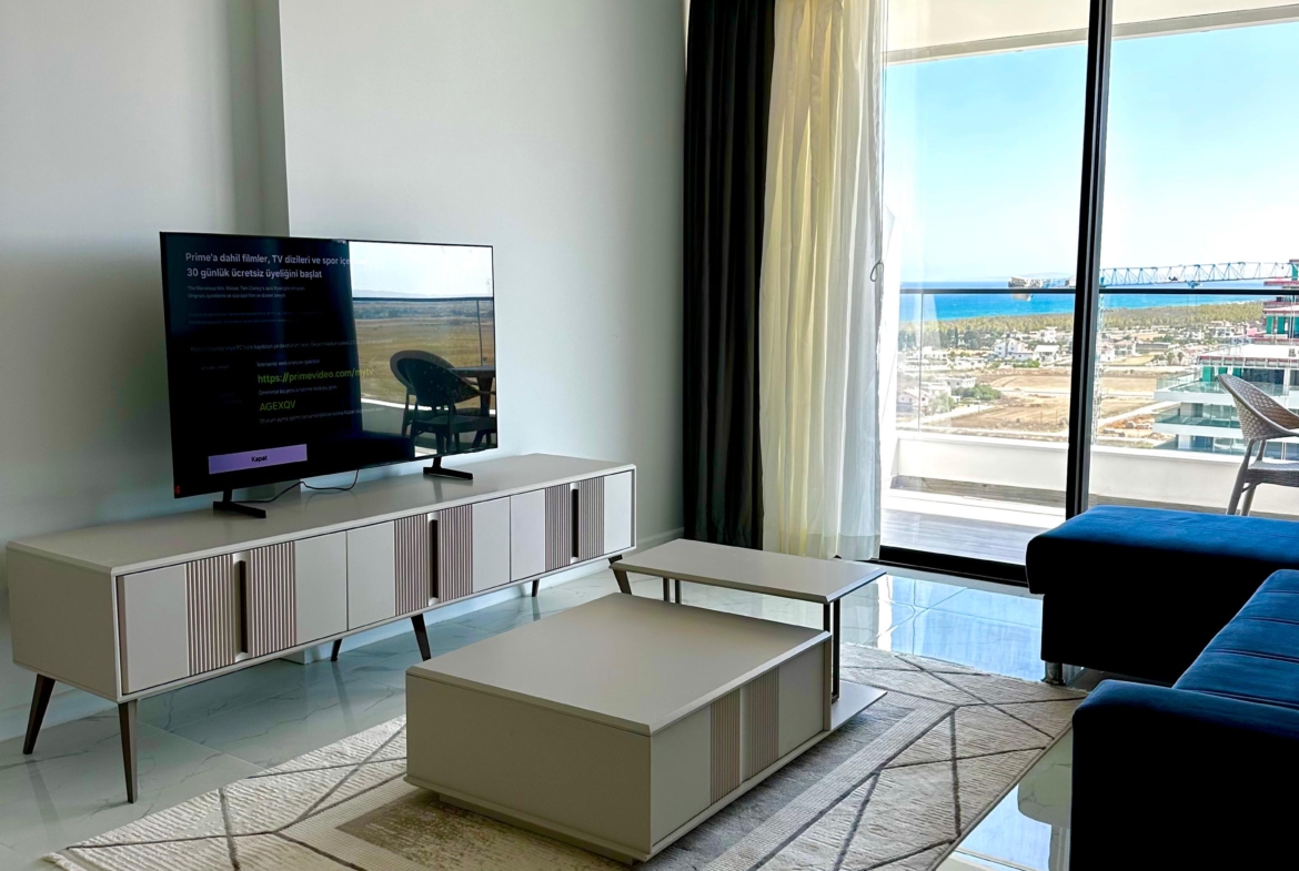 kıbrıs Alçak bir konsol üzerinde bir TV, desenli bir halı üzerinde bir sehpa, koyu renkli bir kanepe ve dışarıda deniz ve gökyüzü manzarası sunan perdeli büyük pencerelere sahip modern oturma odası. Mekan şık ve minimal bir dekorla aydınlıktır. aysan_giti