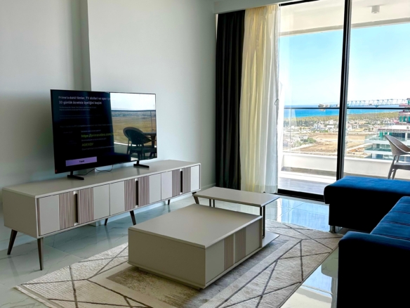 kıbrıs Alçak bir konsol üzerinde bir TV, desenli bir halı üzerinde bir sehpa, koyu renkli bir kanepe ve dışarıda deniz ve gökyüzü manzarası sunan perdeli büyük pencerelere sahip modern oturma odası. Mekan şık ve minimal bir dekorla aydınlıktır. aysan_giti