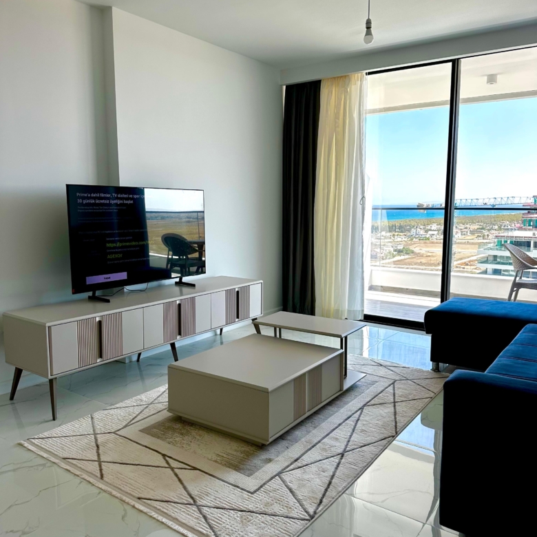 kıbrıs Alçak bir konsol üzerinde bir TV, desenli bir halı üzerinde bir sehpa, koyu renkli bir kanepe ve dışarıda deniz ve gökyüzü manzarası sunan perdeli büyük pencerelere sahip modern oturma odası. Mekan şık ve minimal bir dekorla aydınlıktır. aysan_giti