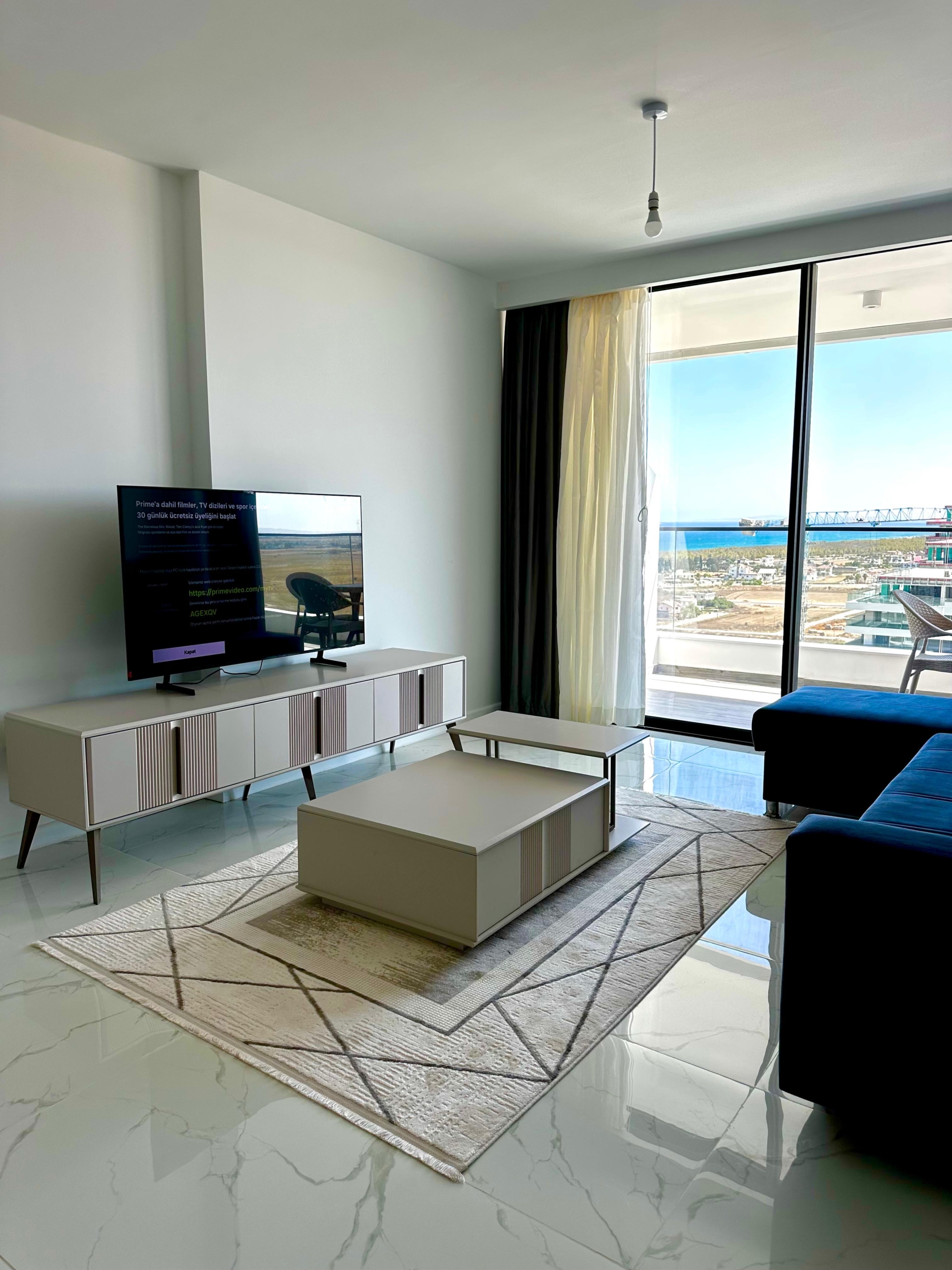 kıbrıs Alçak bir konsol üzerinde bir TV, desenli bir halı üzerinde bir sehpa, koyu renkli bir kanepe ve dışarıda deniz ve gökyüzü manzarası sunan perdeli büyük pencerelere sahip modern oturma odası. Mekan şık ve minimal bir dekorla aydınlıktır. aysan_giti