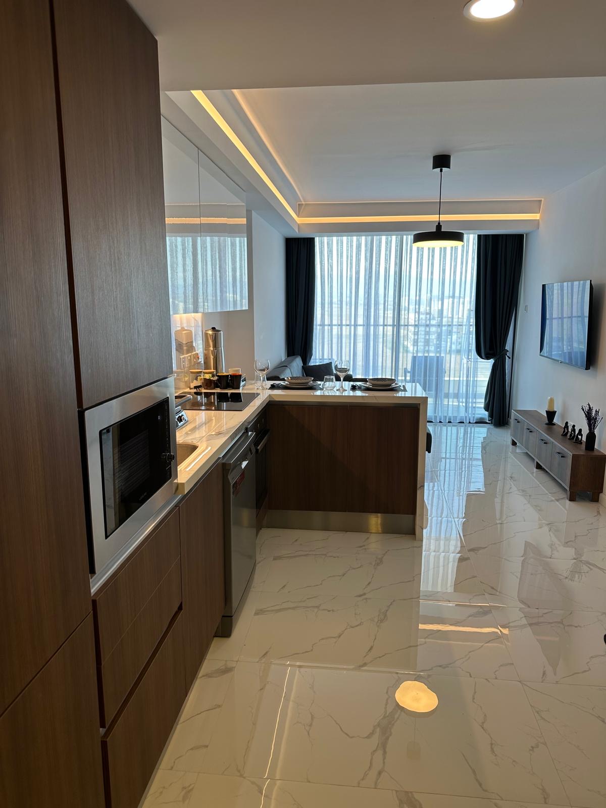 kıbrıs Ahşap dolaplar, ankastre mikrodalga fırın ve beyaz mermer zemin içeren bir mutfağa sahip modern daire iç mekanı. İdeal Kuzey Kıbrıs gayrimenkul seçimi, yemek masası, kanepe, TV ve arka planda tül perdeli büyük pencereler bulunan açık yaşam alanı. aysan_giti