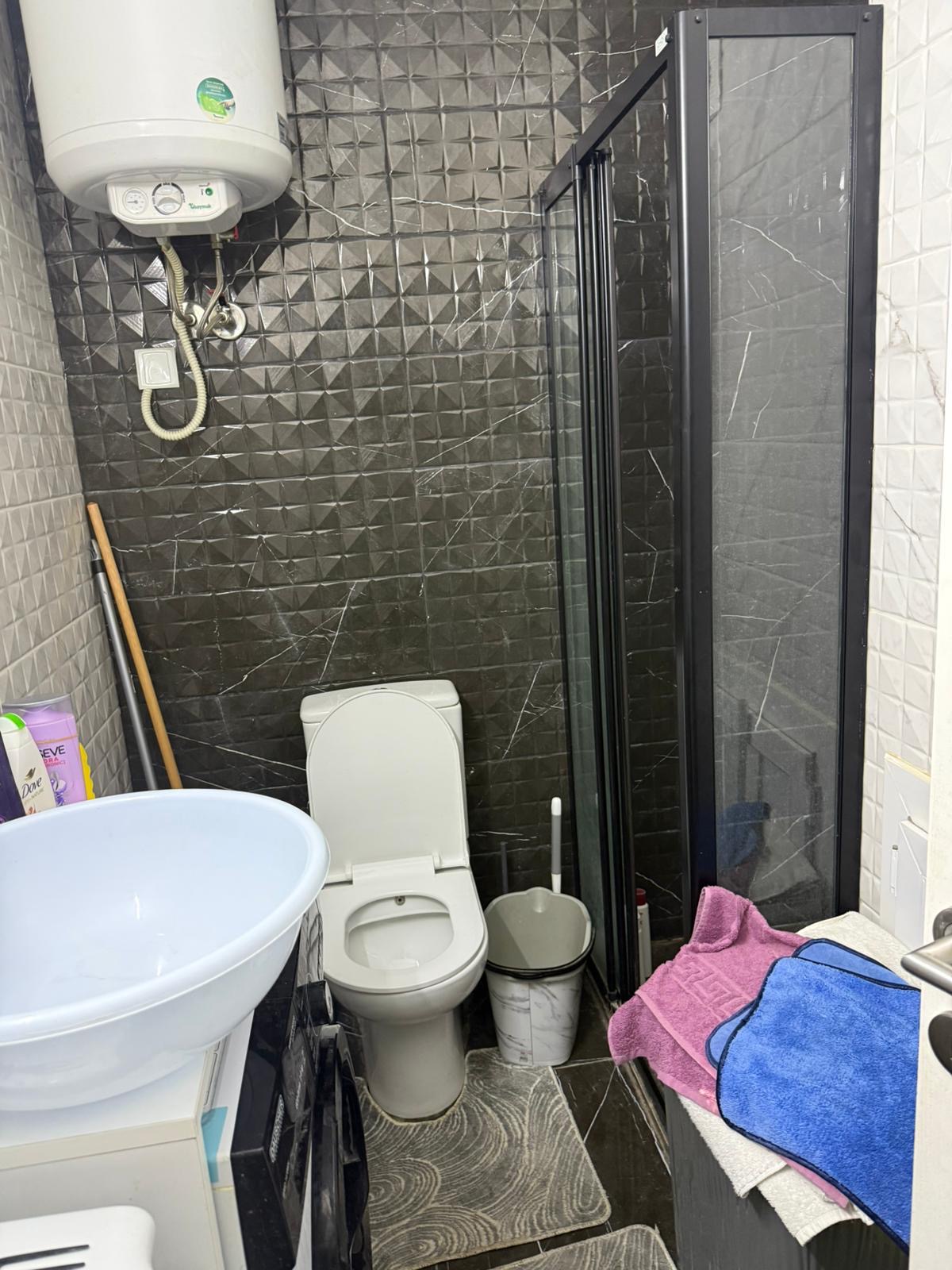 kıbrıs Siyah dokulu duvarları olan küçük bir banyo, bir tuvalet, yuvarlak bir lavabo, cam kapılı bir duş, tuvaletin üzerinde bir su ısıtıcı, bir süpürge, bir paspas kovası ve yakındaki bir yüzeyde renkli havlular. aysan_giti
