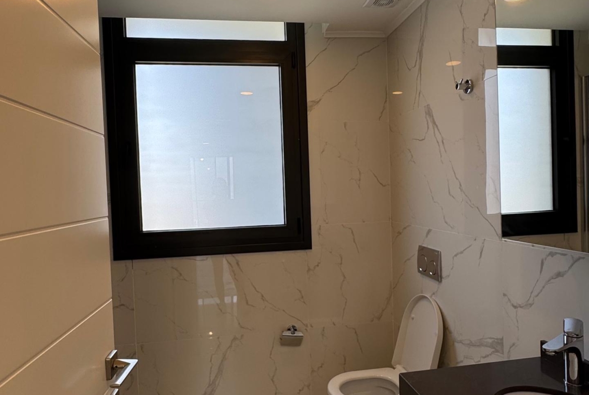 kıbrıs Mermer zemin ve duvarlar, duvara monte tuvalet, koyu renkli tezgah üstü lavabo, geniş buzlu pencere, gömme ışıklar ve kısmen açık beyaz kapıya sahip modern banyo, İskele Long Beach kiralık daire arayanlar için mükemmeldir. aysan_giti