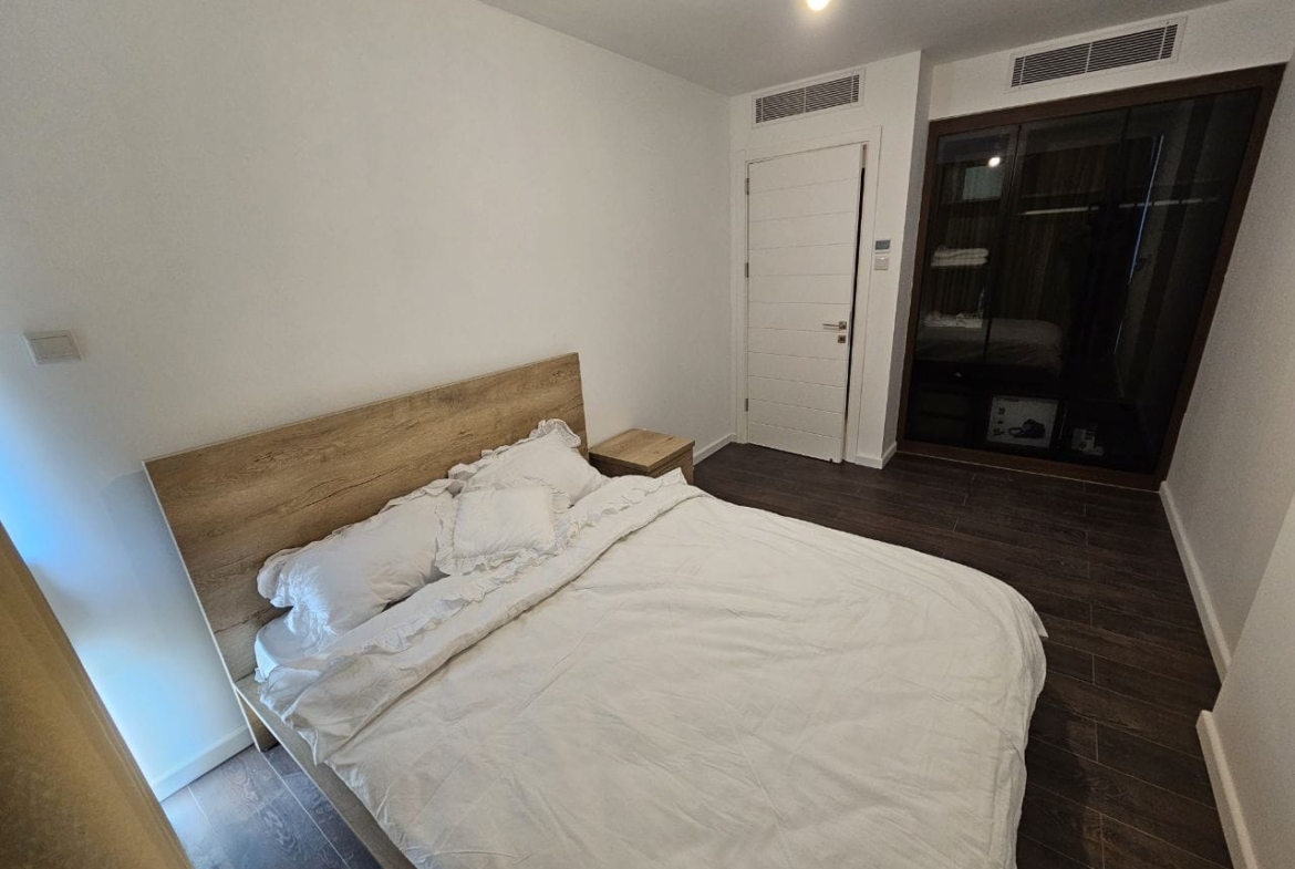 kıbrıs Beyaz nevresimlerle kaplı çift kişilik bir yatak, ahşap bir yatak başlığı ve komodin, koyu ahşap zemin, beyaz duvarlar, kapalı beyaz bir kapı ve büyük koyu çerçeveli pencere veya cam kapı içeren sade bir yatak odası. aysan_giti