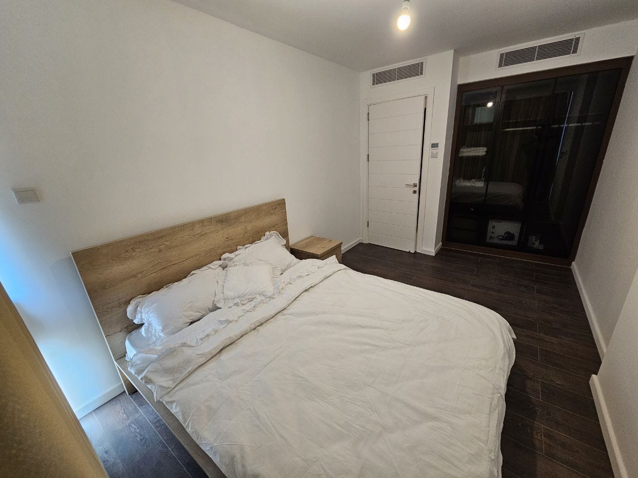 kıbrıs Beyaz nevresimlerle kaplı çift kişilik bir yatak, ahşap bir yatak başlığı ve komodin, koyu ahşap zemin, beyaz duvarlar, kapalı beyaz bir kapı ve büyük koyu çerçeveli pencere veya cam kapı içeren sade bir yatak odası. aysan_giti