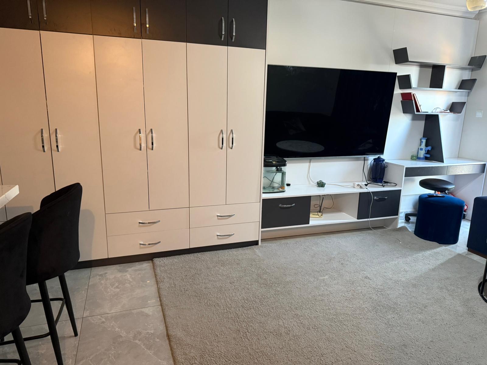 kıbrıs Bej dolaplar, beyaz bir TV sehpası üzerinde büyük bir düz ekran TV, duvarda raflar, iki siyah sandalye ve karo zemin üzerinde açık gri bir halı bulunan bir oturma odası. aysan_giti