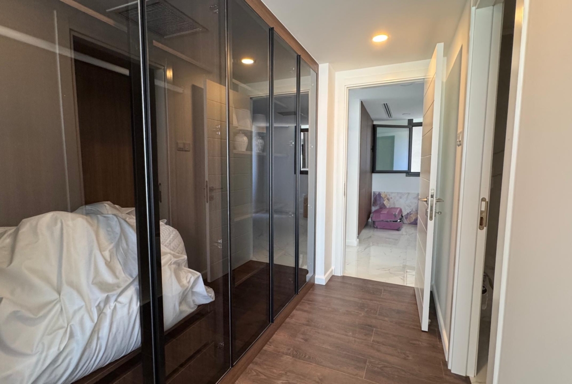 kıbrıs Grand Sapphire Resort & Residence Kıbrıs'ın koridorunda ahşap zemin, solda cam kapılı gardıroplar, beyaz duvarlar ve beyaz karo zeminler ile mor bir kanepeyi ortaya çıkaran açık bir kapı bulunmaktadır. Beyaz yatak takımları gardırop alanının içine yığılmıştır. aysan_giti