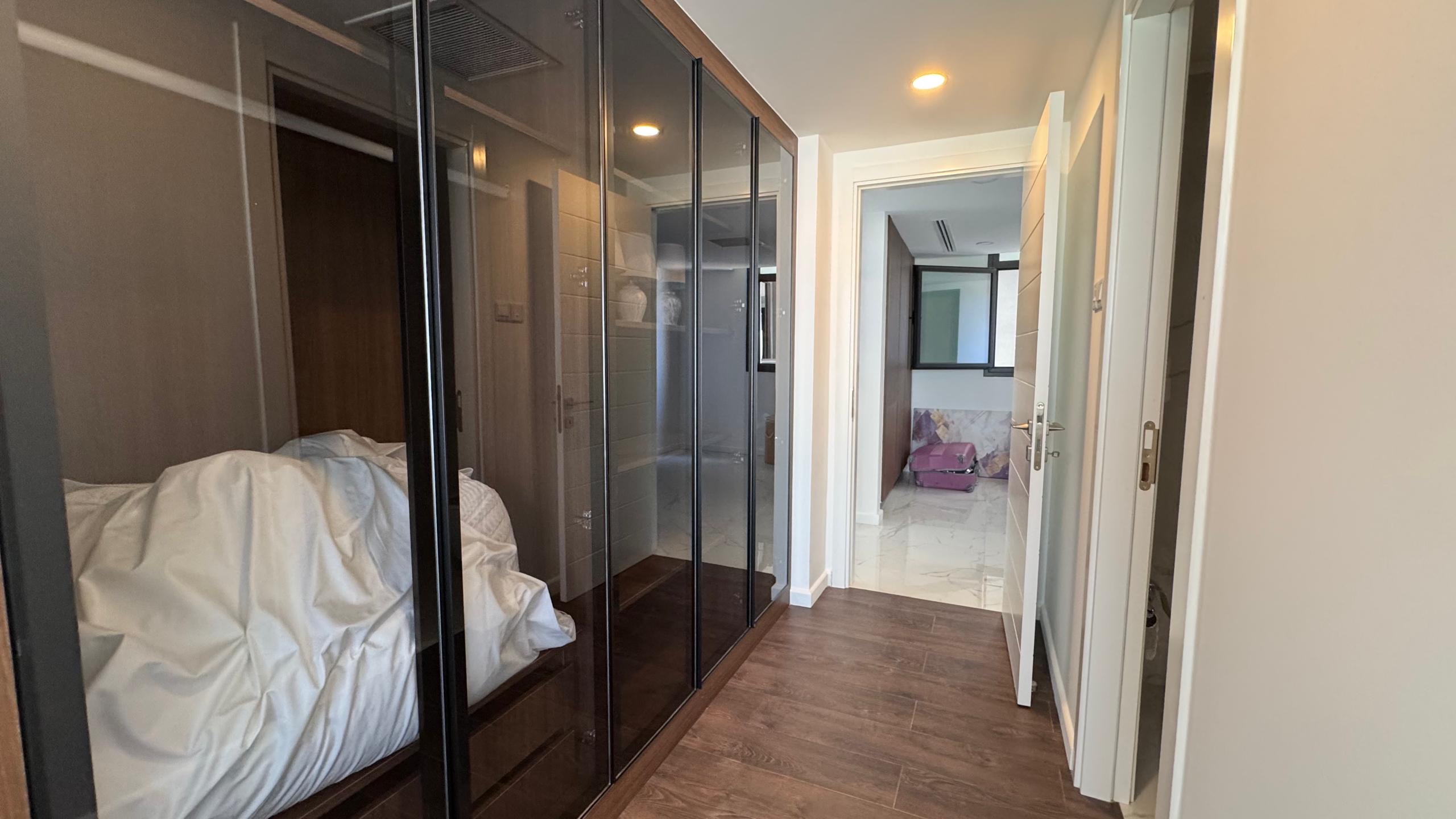 kıbrıs Grand Sapphire Resort & Residence Kıbrıs'ın koridorunda ahşap zemin, solda cam kapılı gardıroplar, beyaz duvarlar ve beyaz karo zeminler ile mor bir kanepeyi ortaya çıkaran açık bir kapı bulunmaktadır. Beyaz yatak takımları gardırop alanının içine yığılmıştır. aysan_giti