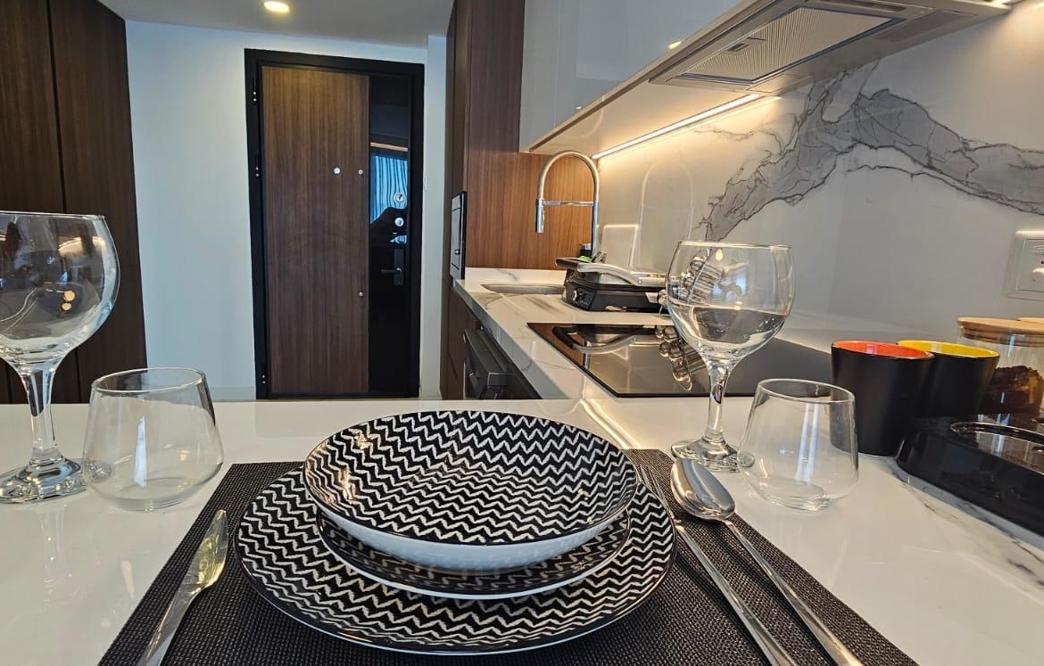 kıbrıs Siyah beyaz desenli tabaklar, bardaklar ve çatal bıçak takımlarıyla modern bir mutfak yemek düzeni, şık bir Kuzey Kıbrıs satılık daire içindeki bir masadan görülüyor. Gri damarlı beyaz tezgah ve şık bir set üstü ocak görünümü tamamlıyor. aysan_giti