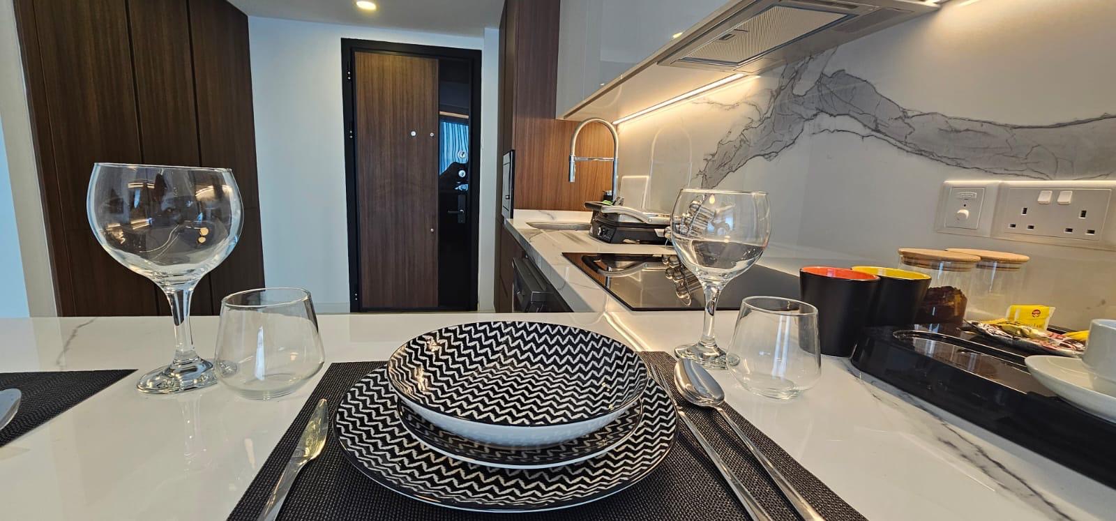 kıbrıs Siyah beyaz desenli tabaklar, bardaklar ve çatal bıçak takımlarıyla modern bir mutfak yemek düzeni, şık bir Kuzey Kıbrıs satılık daire içindeki bir masadan görülüyor. Gri damarlı beyaz tezgah ve şık bir set üstü ocak görünümü tamamlıyor. aysan_giti